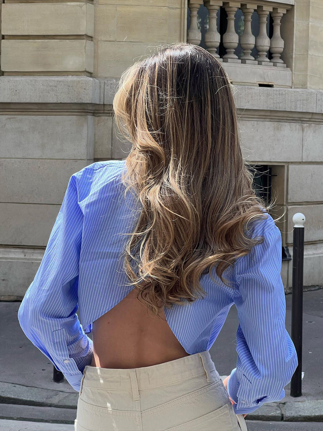 Vue arrière du modèle portant une chemise rayée bleu et blanc à manches longues, avec un dos ouvert pour un look audacieux et élégant. Elle l’associe à un pantalon beige taille haute, créant un style chic et moderne.