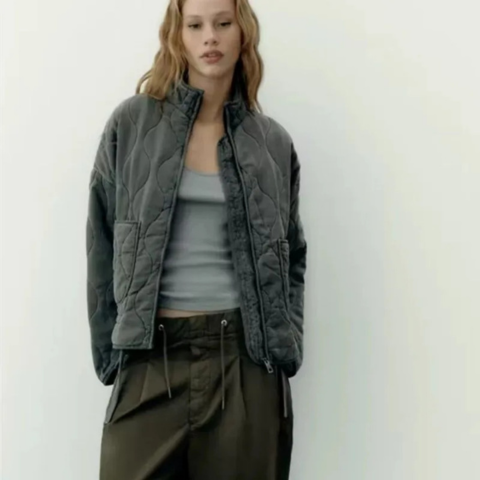 Mannequin portant une veste matelassée grise à col montant et fermeture zippée, avec motif matelassage ondulé, associée à un débardeur clair et un pantalon kaki.