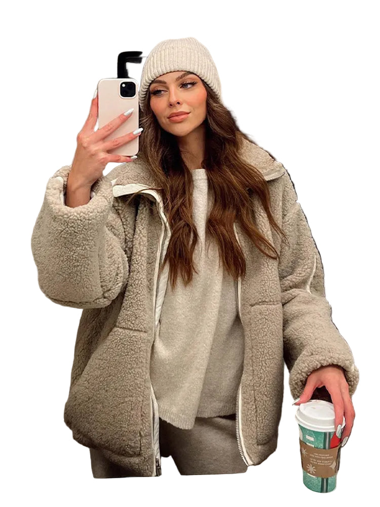 Femme portant une veste en teddy beige, avec un col haut et une coupe décontractée, posant en selfie dans un miroir. Elle porte un pull beige et un pantalon assorti, avec un bonnet tricoté beige. Elle tient une tasse de café dans ses mains, affichant un look cosy et élégant.