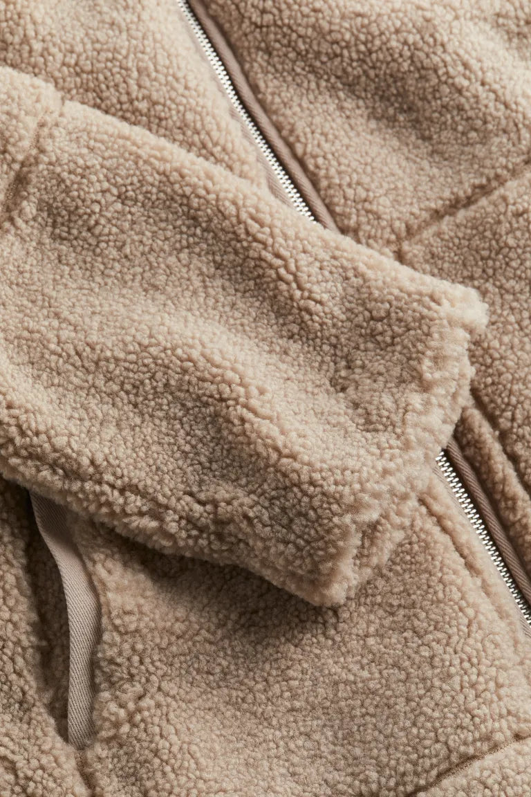 Un gros plan sur la manche d'une veste teddy marron révèle la texture douce et moelleuse du tissu. Les poignets sont finis dans un tissu beige assorti, avec une fermeture éclair argentée sur le côté, soulignant son allure à la fois élégante et confortable.