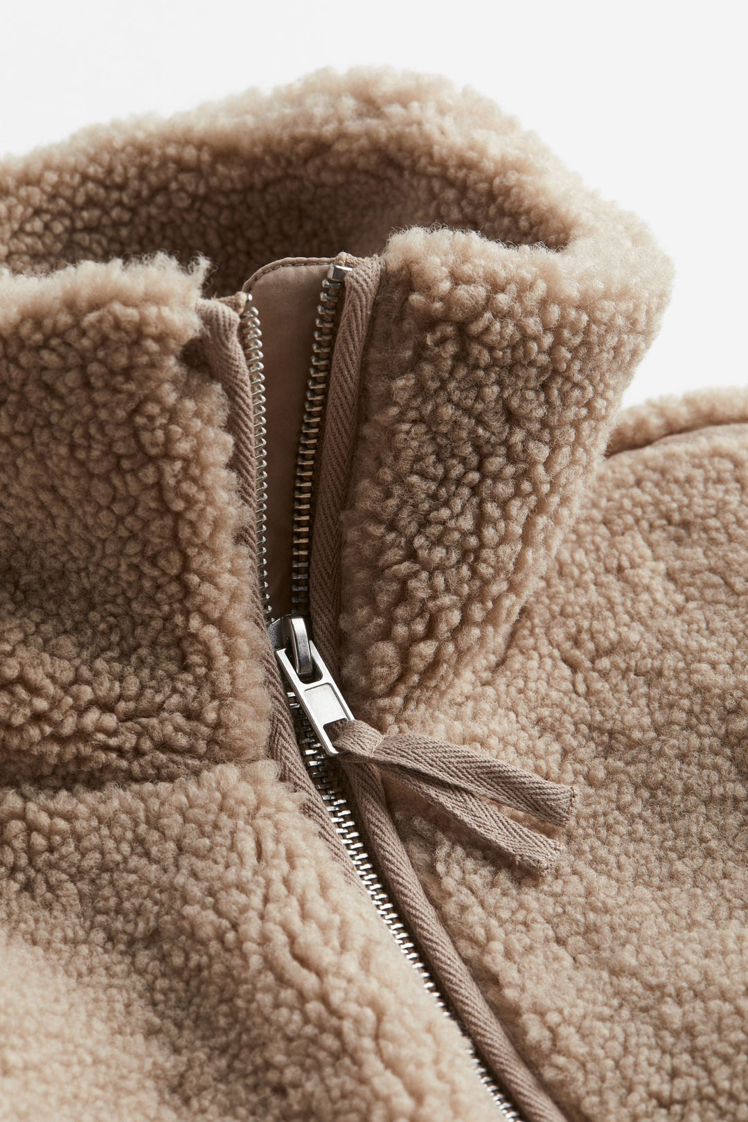 Gros plan sur la fermeture éclair d'une veste teddy marron, avec des tirettes en tissu beige assorties. La texture douce et duveteuse du tissu est mise en valeur autour de la fermeture éclair, créant un style chaleureux et soigné.