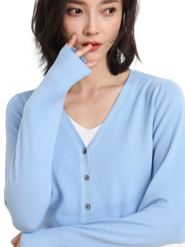 Femme portant un cardigan col V bleu pastel boutonné, superposé à un haut blanc, posant de profil avec une main près du visage, sur un fond uni clair.