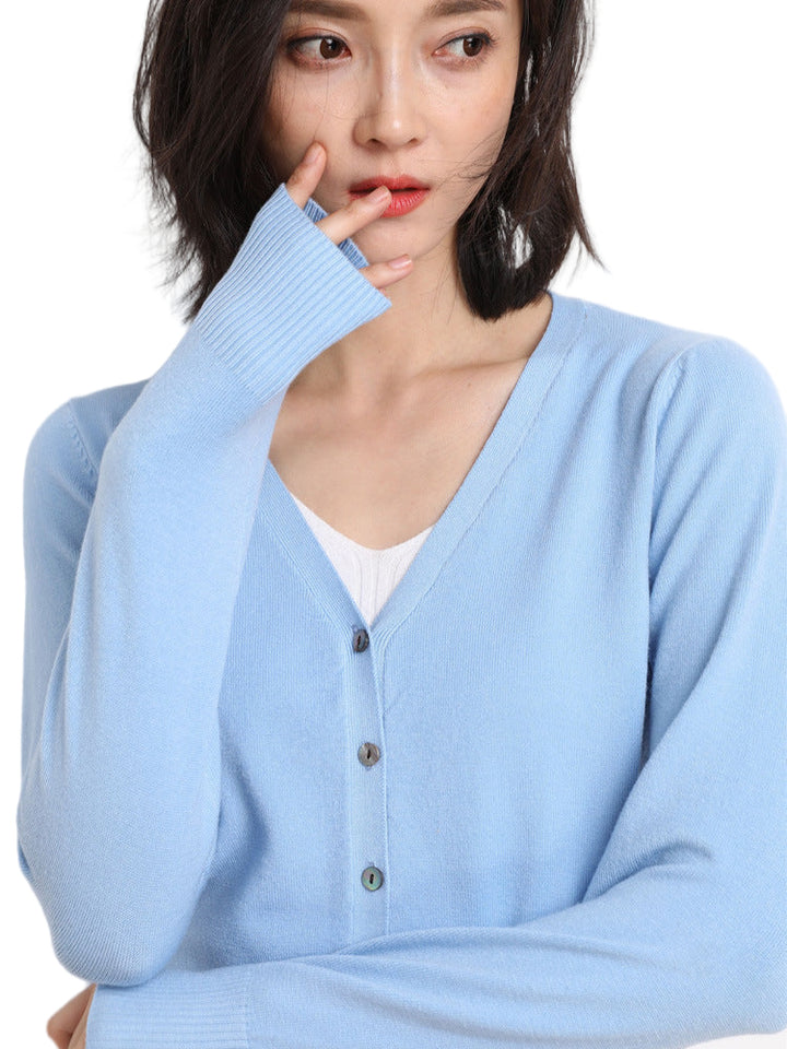 Femme portant un cardigan col V bleu pastel boutonné, superposé à un haut blanc, posant de profil avec une main près du visage, sur un fond uni clair.