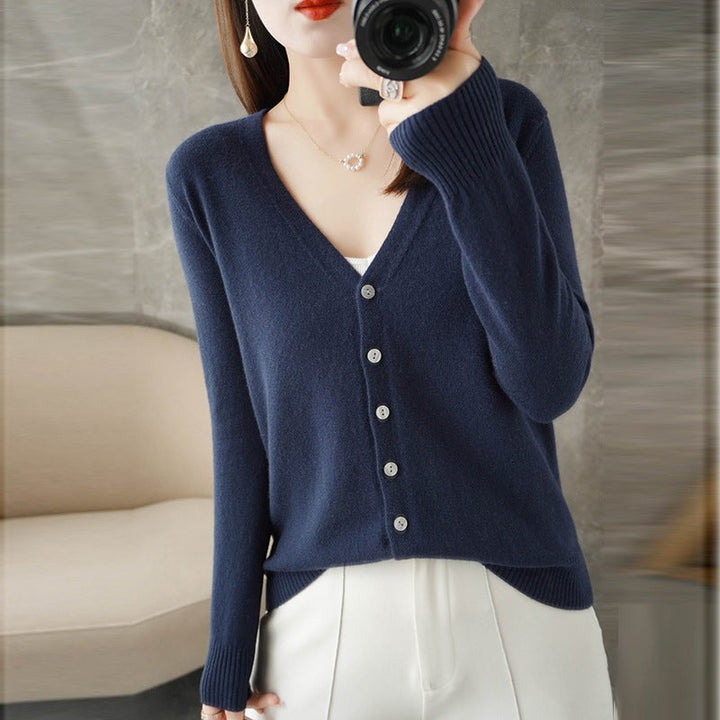 Femme portant un cardigan bleu marine à col V boutonné, superposé à un haut blanc, assorti à un pantalon clair, et prenant une photo avec un appareil photo dans un intérieur moderne.