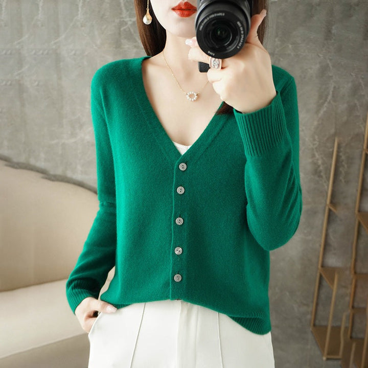Femme portant un cardigan vert à col V boutonné, superposé à un haut blanc et assorti à un pantalon clair, prenant une photo avec un appareil photo dans un intérieur moderne.