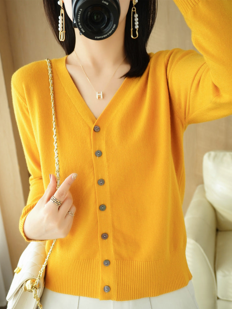 Femme portant un cardigan jaune à col V boutonné, assorti à un pantalon clair et accompagné d’un sac à chaîne dorée, prenant une photo avec un appareil photo dans un intérieur lumineux.