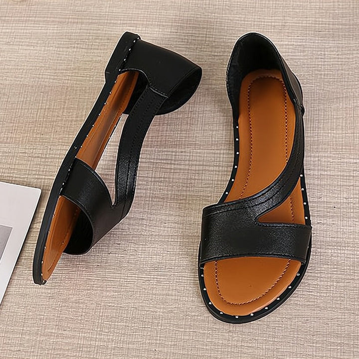 Sandales plates femme Capucine tendance noires, bout ouvert, bride large asymétrique avec découpes, semelle camel et bord clouté, présentées en vue de dessus sur fond clair.