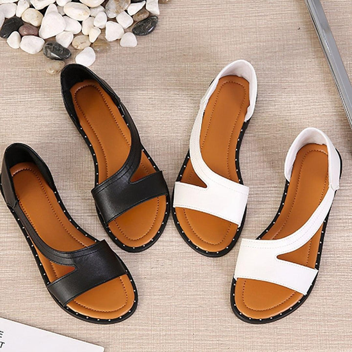 Sandales plates femme Capucine tendance en noir et blanc, bout ouvert, bride large asymétrique avec découpes, semelle camel et contour clouté, présentées en vue de dessus sur fond clair.