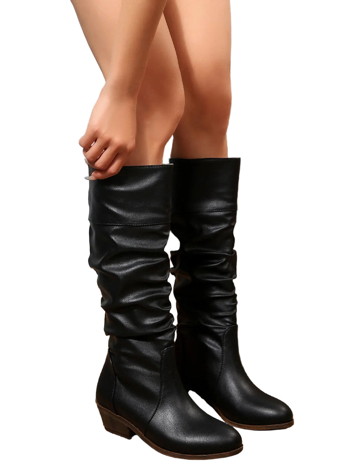 Femme ajustant des bottes mi-mollet noires en similicuir, dotées d’un talon bloc bas et d’un design plissé, posant dans un décor rustique avec un tapis en jute, un cactus et des éléments en bois.