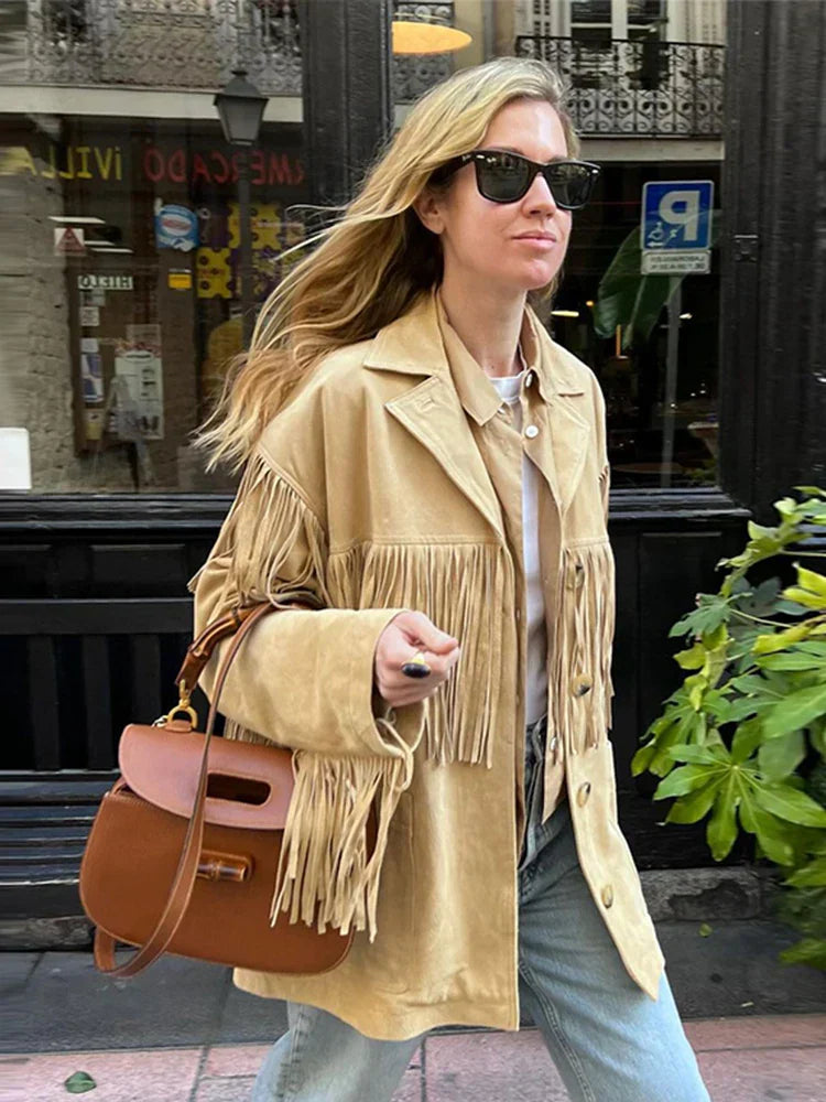 Veste à franges pour femme, modèle Charlène. Veste en suède beige avec des franges sur les manches et le bas, portée avec une chemise beige et un sac à main en cuir. Un look tendance et élégant, parfait pour un style moderne et décontracté.