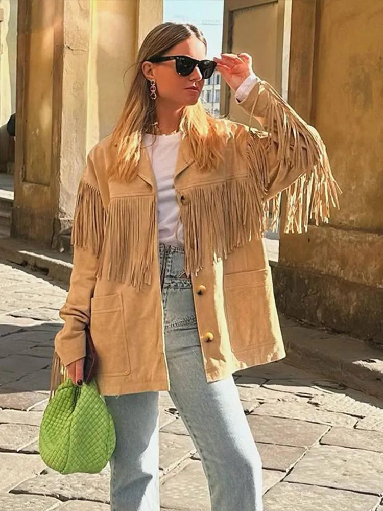 Veste à franges pour femme, modèle Charlène. Veste beige à franges sur les manches, portée avec un t-shirt blanc, un jean taille haute et un sac à main vert. Look tendance et décontracté, parfait pour les journées ensoleillées.