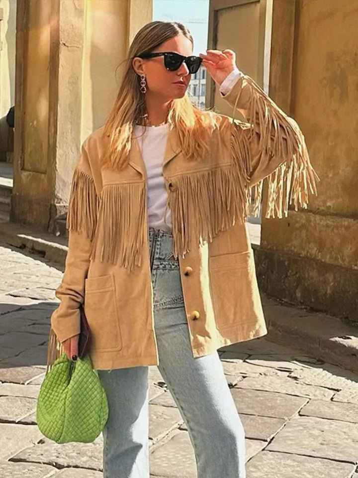 Veste à franges pour femme, modèle Charlène. Veste beige à franges sur les manches, portée avec un t-shirt blanc, un jean taille haute et un sac à main vert. Look tendance et décontracté, parfait pour les journées ensoleillées.