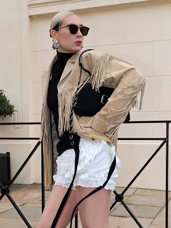 Veste à franges pour femme, modèle Charlène. Veste beige à franges sur les manches, portée avec un pull noir, un short blanc à volants et un sac à main noir. Accessoires modernes avec lunettes de soleil et boucles d'oreilles, pour un look chic et tendance.