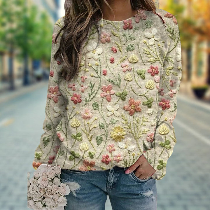 Pull beige à manches longues avec broderies florales colorées en relief, porté avec un jean, sur fond urbain flouté.