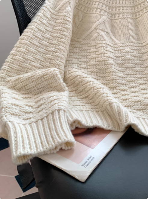 Gros plan sur un pull blanc en maille, mettant en valeur ses poignets côtelés et ses motifs texturés, posé sur une chaise avec un magazine en dessous, dans un intérieur moderne.