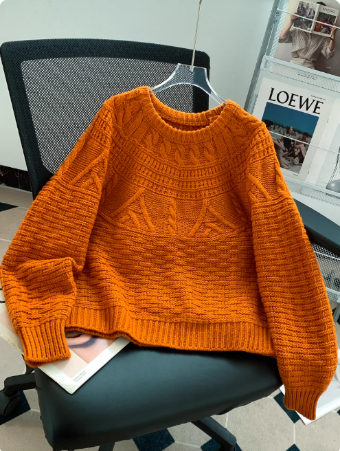 Pull en maille orange vif à col rond, avec motifs texturés et finitions côtelées, présenté sur un cintre et posé sur une chaise de bureau dans un intérieur moderne avec un porte-magazines en arrière-plan.