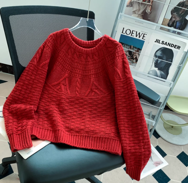 Pull en maille rouge à col rond, avec motifs texturés et finitions côtelées, présenté sur un cintre et posé sur une chaise de bureau dans un intérieur moderne avec un porte-magazines en arrière-plan.