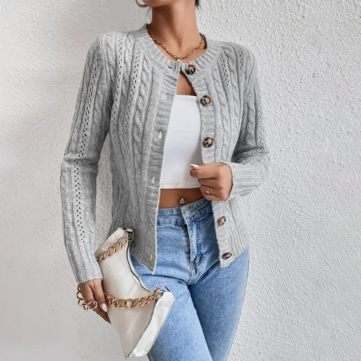 Femme portant un cardigan en maille torsadée gris clair, légèrement ouvert sur un haut court blanc, assorti à un jean taille haute bleu clair et tenant une pochette blanche à chaîne dorée.