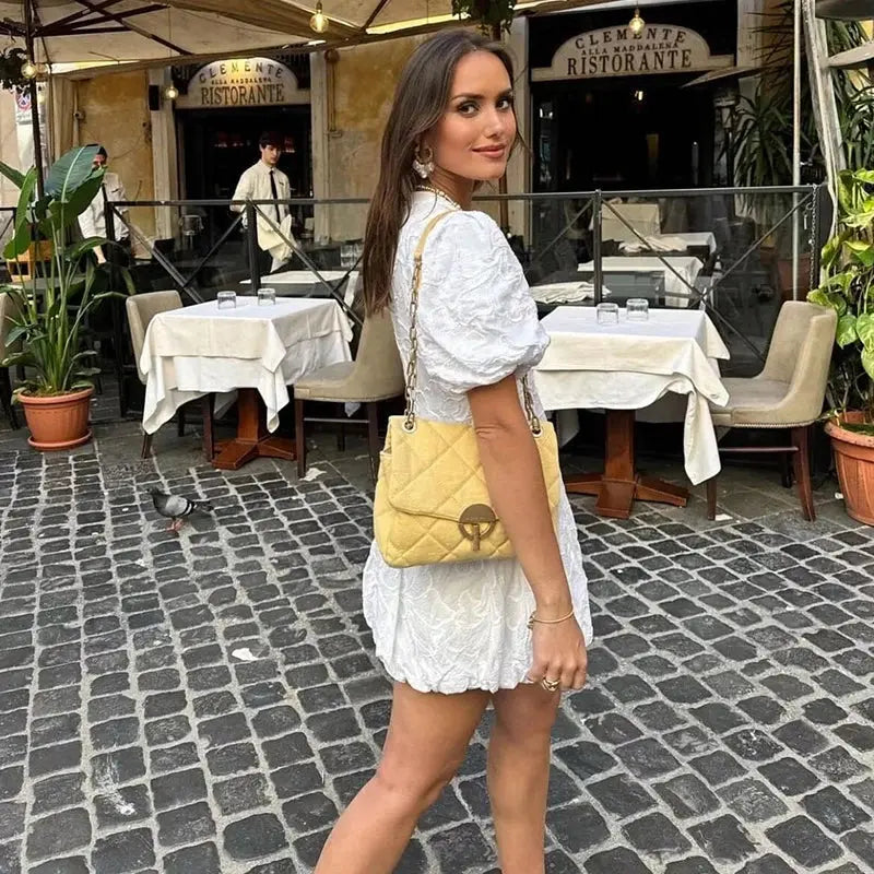Femme portant une mini-robe blanche à imprimé floral, style moderne, avec des manches bouffantes et un design élégant. Elle porte un sac à main jaune en bandoulière et pose dans une rue pavée à côté d'un restaurant. Idéale pour un look chic et décontracté.