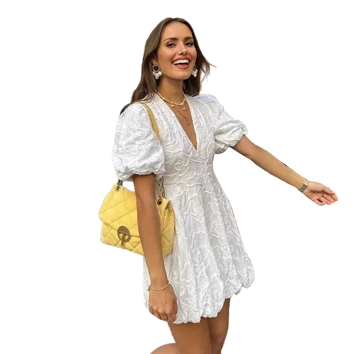 Femme souriante portant une mini-robe blanche à imprimé floral et à manches bouffantes, avec un décolleté en V. Elle porte des bijoux élégants et un sac à main jaune, tout en posant dans une rue pavée près d'un restaurant. Une tenue moderne et chic, parfaite pour un look estival.
