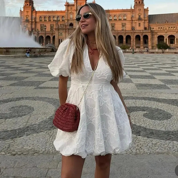 Femme portant une mini-robe blanche à imprimé floral avec des manches bouffantes, posant en plein air devant un magnifique bâtiment. Elle porte des lunettes de soleil, des bijoux élégants et un sac à main rouge. Un look moderne et estival, parfait pour les journées ensoleillées.