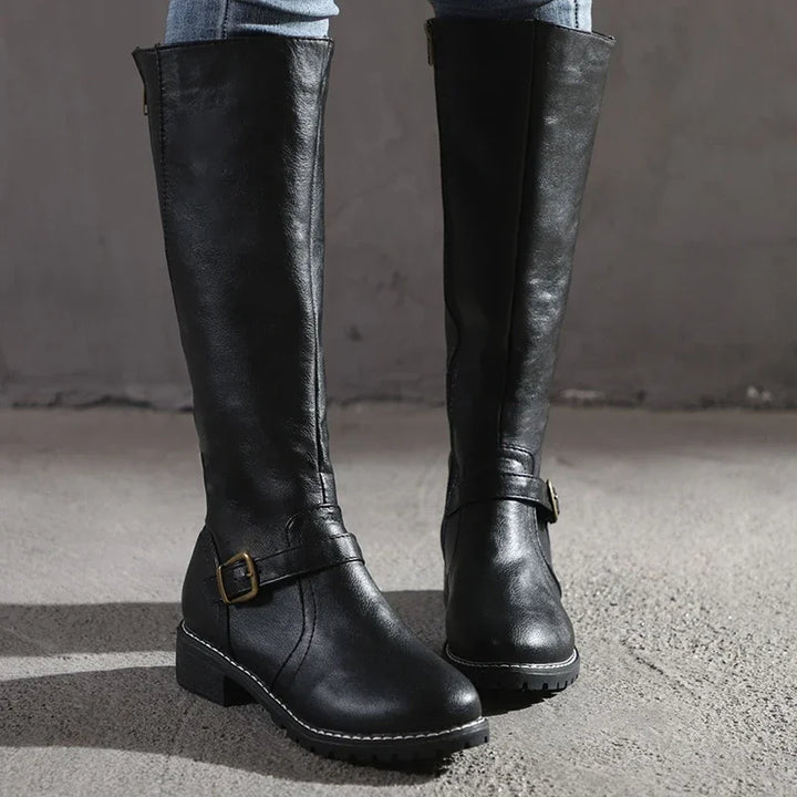 Paire de bottes hautes noires en cuir synthétique, dotées d’une fermeture éclair latérale et d’une bride décorative avec boucle métallique dorée à la cheville. Leur semelle crantée et leur talon bas assurent confort et stabilité pour un usage quotidien. Ces bottes au style sobre et élégant sont idéales pour l’automne et l’hiver, parfaites avec un jean ou une robe pour un look moderne et raffiné.