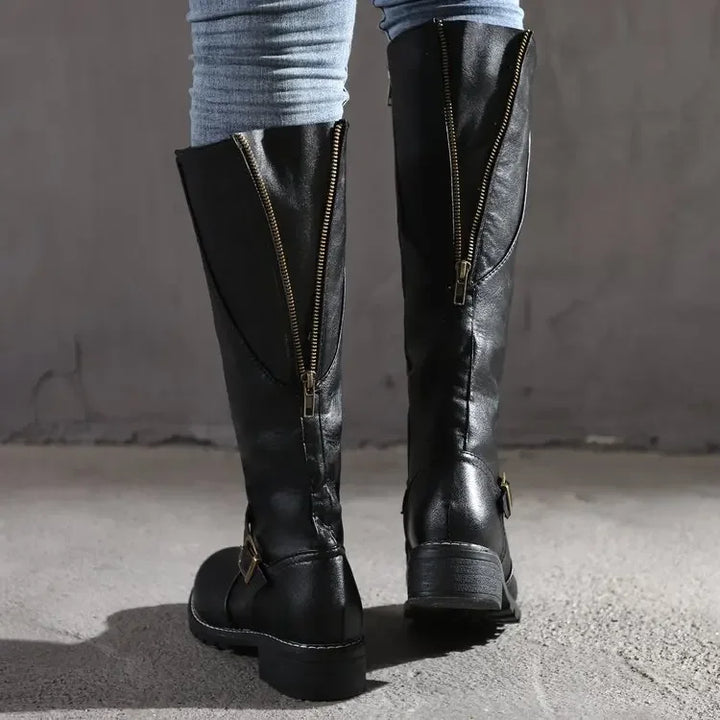 Vue arrière d’une paire de bottes hautes noires en cuir synthétique, dotées de fermetures éclair apparentes à l’arrière et de brides décoratives avec boucles dorées au niveau des chevilles. Les bottes présentent un talon bas et une semelle antidérapante, offrant à la fois confort et style. Leur design moderne et pratique les rend parfaites pour la saison automne-hiver.