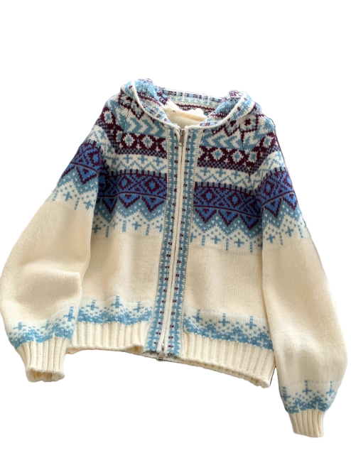 Sweat à capuche blanc en maille zippée, orné de motifs géométriques bleus et bordeaux, présenté sur un cintre et posé sur une chaise de bureau dans un intérieur moderne avec un porte-revues en arrière-plan.