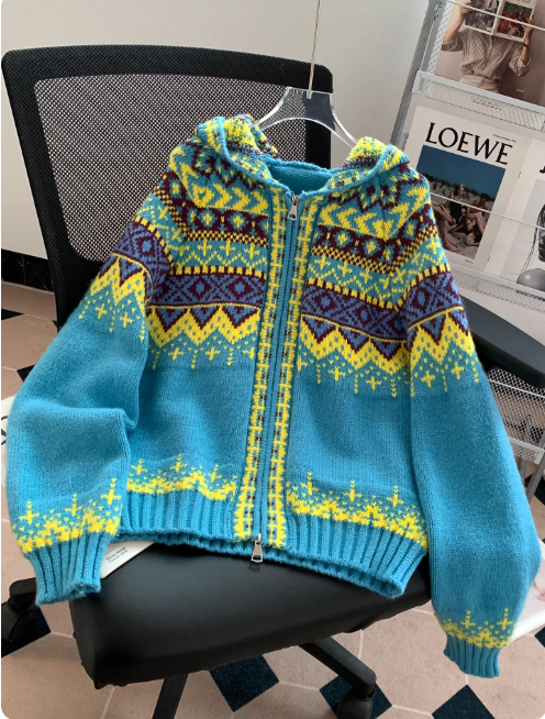 Pull à capuche en maille bleu turquoise avec fermeture éclair, orné de motifs géométriques jaunes, violets et bordeaux, présenté sur un cintre et posé sur une chaise de bureau dans un intérieur moderne avec un porte-magazines en arrière-plan.