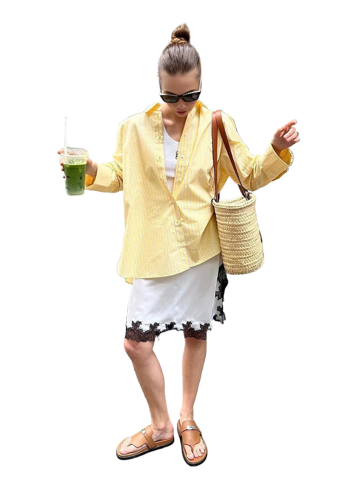Le modèle porte une chemise rayée jaune et blanche à manches longues, ouverte sur un top blanc. Elle associe la chemise à une jupe blanche ornée de dentelle noire et des sandales marron, créant un look estival et élégant. Elle tient un verre de smoothie vert et porte un sac en osier, parfait pour une journée décontractée en ville.