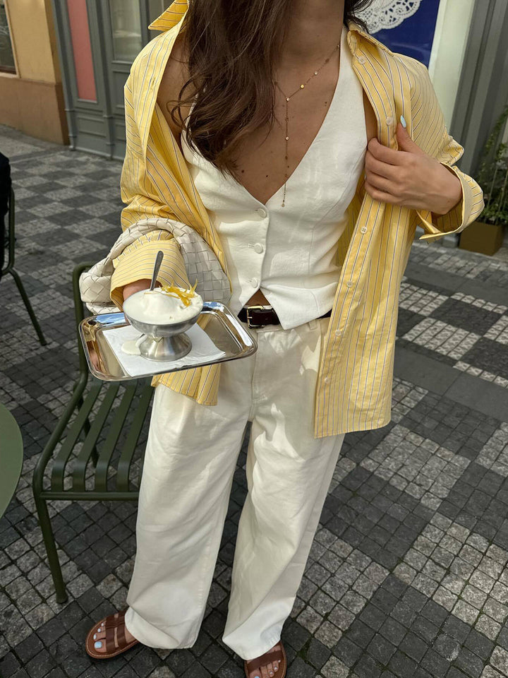 Le modèle porte une chemise rayée jaune et blanche à manches longues, ouverte sur un top blanc, associée à un pantalon blanc fluide. Elle tient un plateau avec un dessert léger, créant une silhouette estivale, chic et décontractée, parfaite pour une sortie en terrasse.