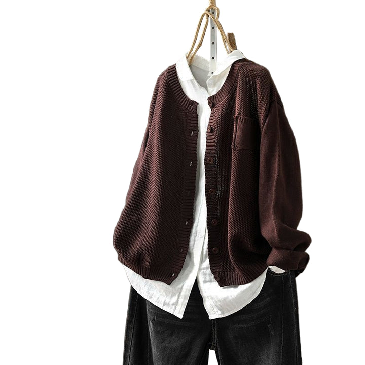 Un cardigan oversize couleur café, suspendu à un cintre, porté sur une chemise blanche boutonnée et associé à un pantalon foncé, est présenté sur un fond en bois foncé.