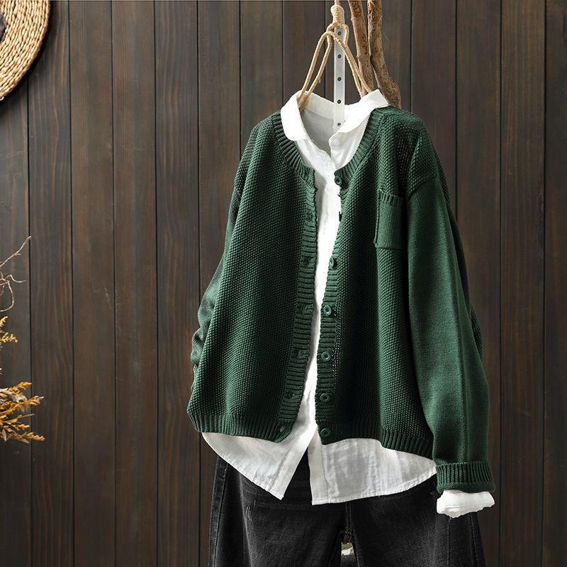 Cardigan oversize vert suspendu sur un cintre, porté sur une chemise blanche ample et assorti à un pantalon foncé, présenté devant un fond en bois sombre.