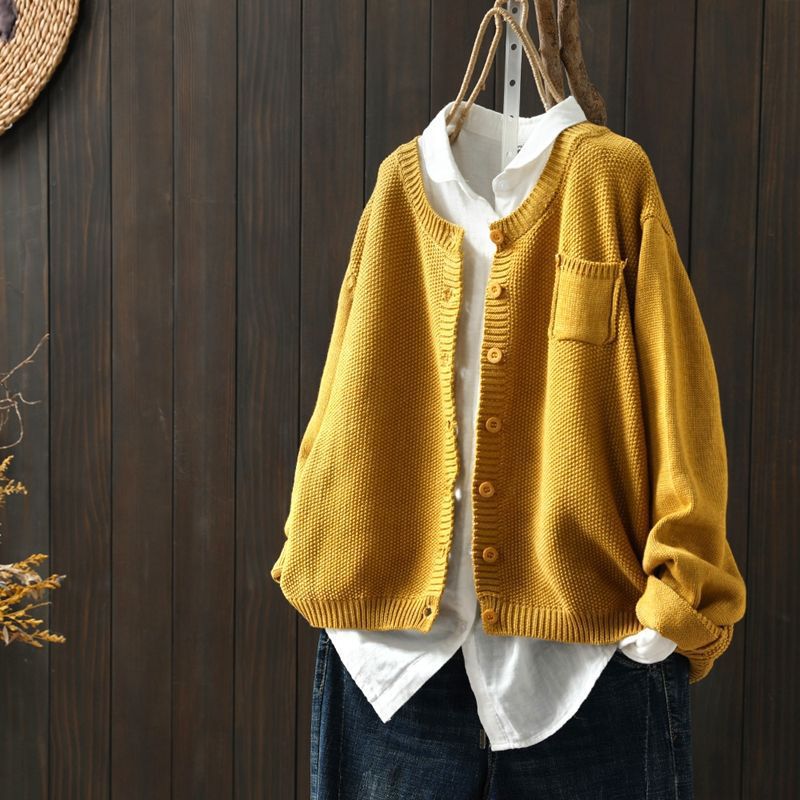Cardigan oversize jaune suspendu sur un cintre, porté sur une chemise blanche ample et assorti à un pantalon en denim foncé, présenté devant un fond en bois sombre.