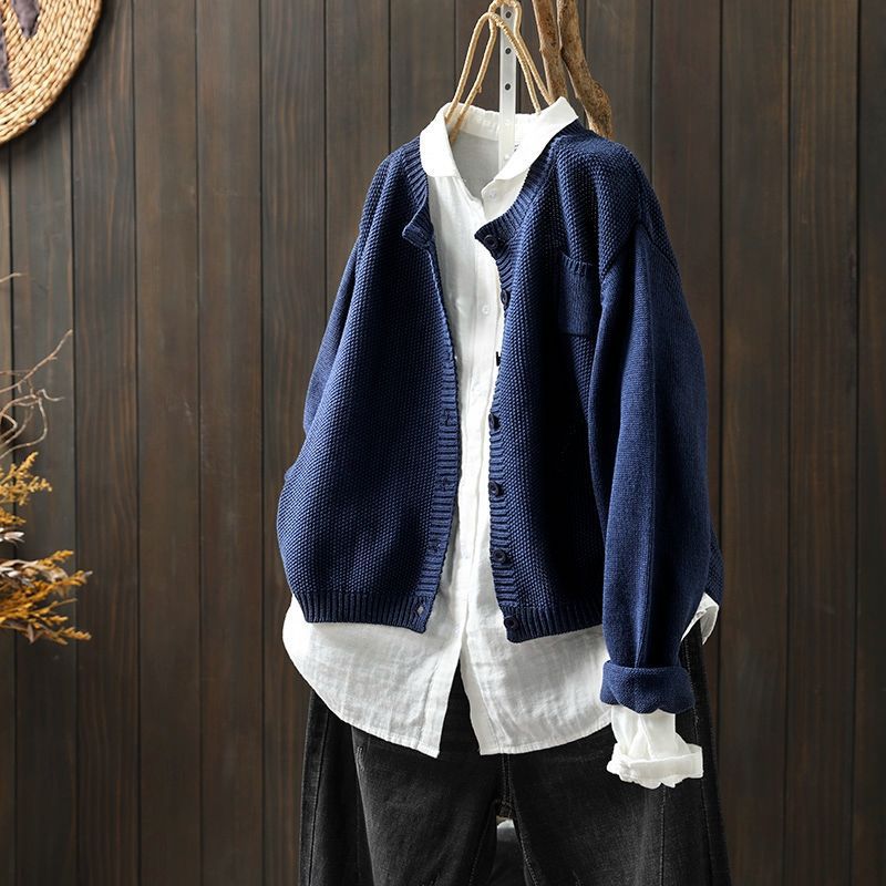 Cardigan oversize bleu marine suspendu sur un cintre, porté sur une chemise blanche ample et assorti à un pantalon foncé, présenté devant un fond en bois sombre.