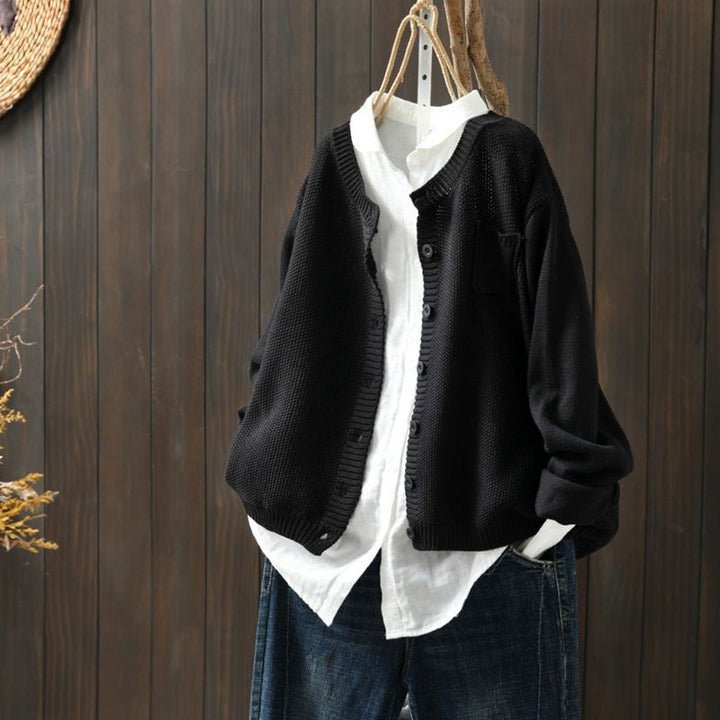 Cardigan oversize noir suspendu sur un cintre, porté sur une chemise blanche ample et assorti à un pantalon en denim bleu, présenté devant un fond en bois sombre.
