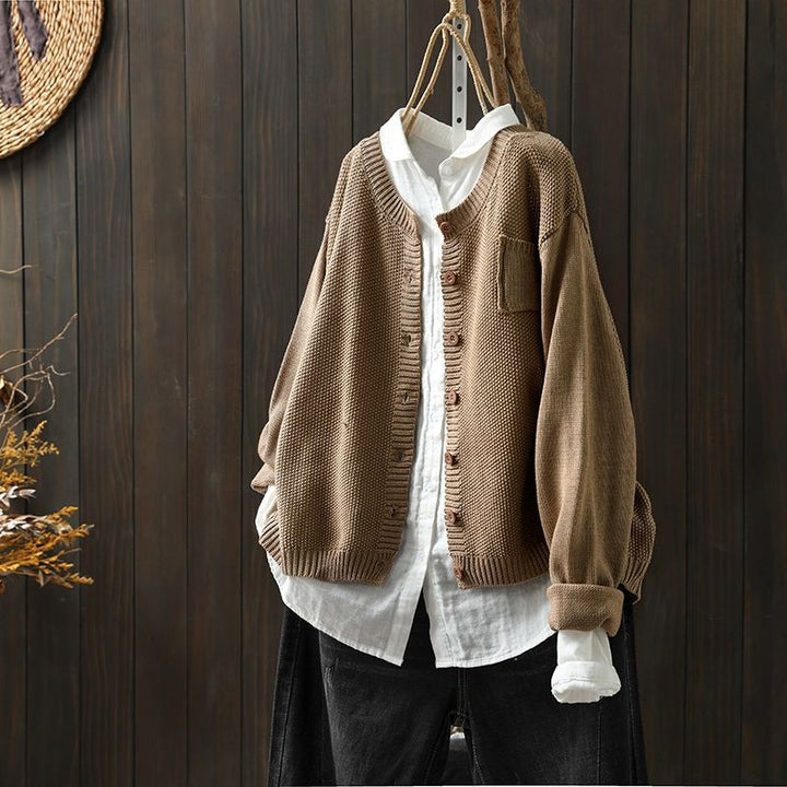 Cardigan oversize kaki suspendu sur un cintre, superposé à une chemise blanche ample et associé à un pantalon foncé, présenté devant un fond en bois sombre.