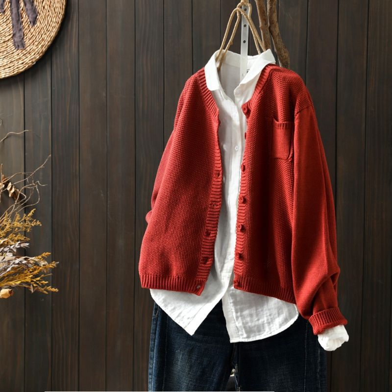 Cardigan oversize rouge suspendu sur un cintre, superposé à une chemise blanche ample et assorti à un pantalon en denim foncé, présenté devant un fond en bois sombre.
