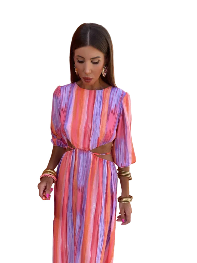 Robe longue aux motifs tie-dye vibrants dans des tons de rose, violet et orange, avec des découpes élégantes sur les côtés. Accessoirisée avec un sac à main rouge et des bijoux dorés, cette robe incarne un style audacieux et moderne, idéale pour les occasions estivales.