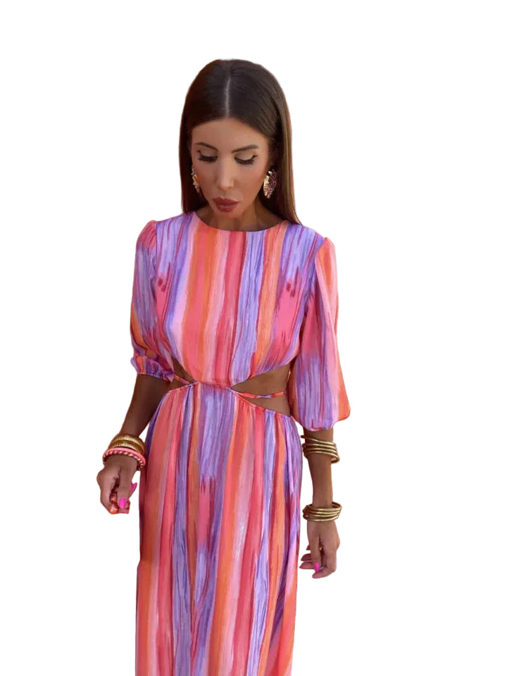 Robe longue aux motifs tie-dye vibrants dans des tons de rose, violet et orange, avec des découpes élégantes sur les côtés. Accessoirisée avec un sac à main rouge et des bijoux dorés, cette robe incarne un style audacieux et moderne, idéale pour les occasions estivales.