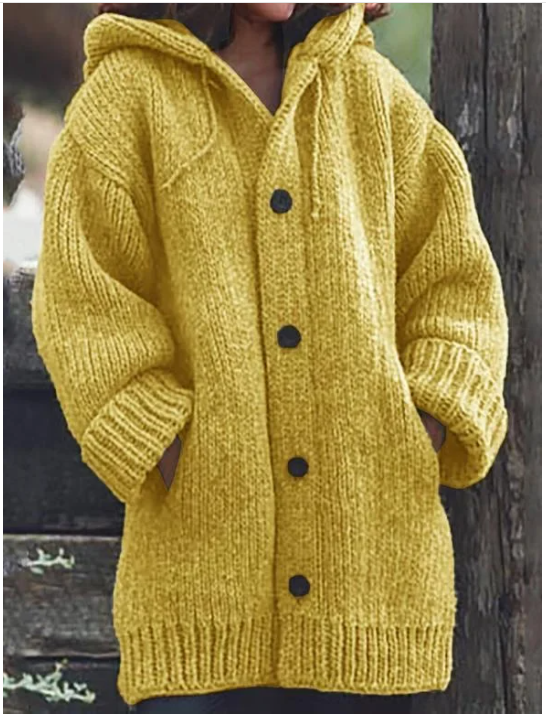 Femme portant un long gilet en maille épaisse jaune moutarde, doté d’une capuche, de manches amples, de poches latérales et de gros boutons foncés, offrant un style hivernal chaud et décontracté.