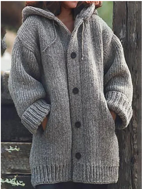 Femme portant un long gilet en maille épaisse gris chiné, avec capuche, manches amples, poches latérales et gros boutons foncés, pour un style hivernal chaud et décontracté.