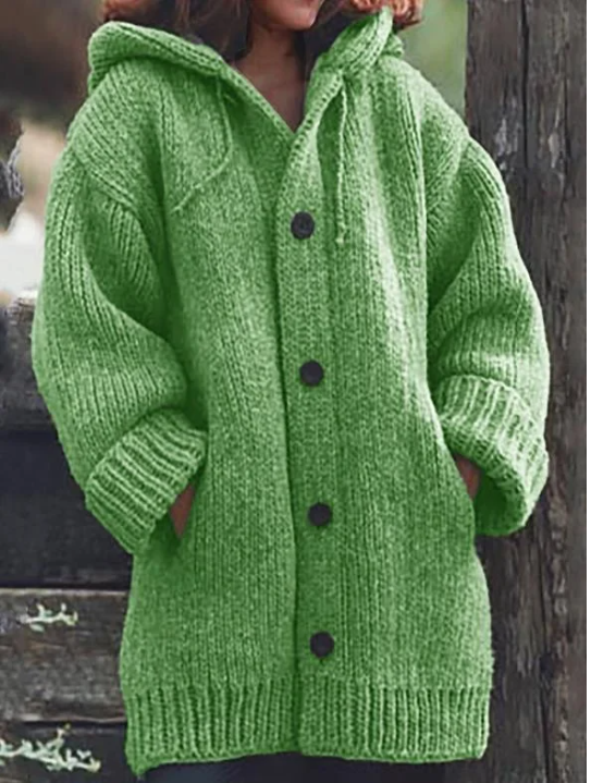 Femme portant un long gilet en maille épaisse vert vif, avec capuche, manches amples, poches latérales et gros boutons foncés, offrant un style hivernal chaud, moderne et décontracté.