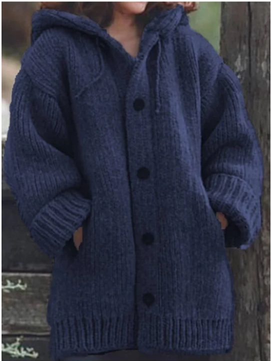 Femme portant un long gilet bleu foncé en maille épaisse avec capuche, manches larges et gros boutons ton sur ton, offrant un look confortable et chaud, idéal pour l'hiver.