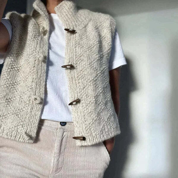 Femme portant un gilet en maille texturée de couleur blanche, ouvert sur un t-shirt blanc et associé à un pantalon clair, avec des boutons en bois apparents sur le devant.