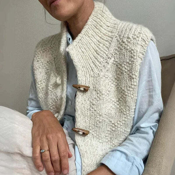 Femme assise portant un gilet en maille texturée de couleur blanche, légèrement ouvert sur une chemise claire, avec des boutons en bois visibles sur le devant.