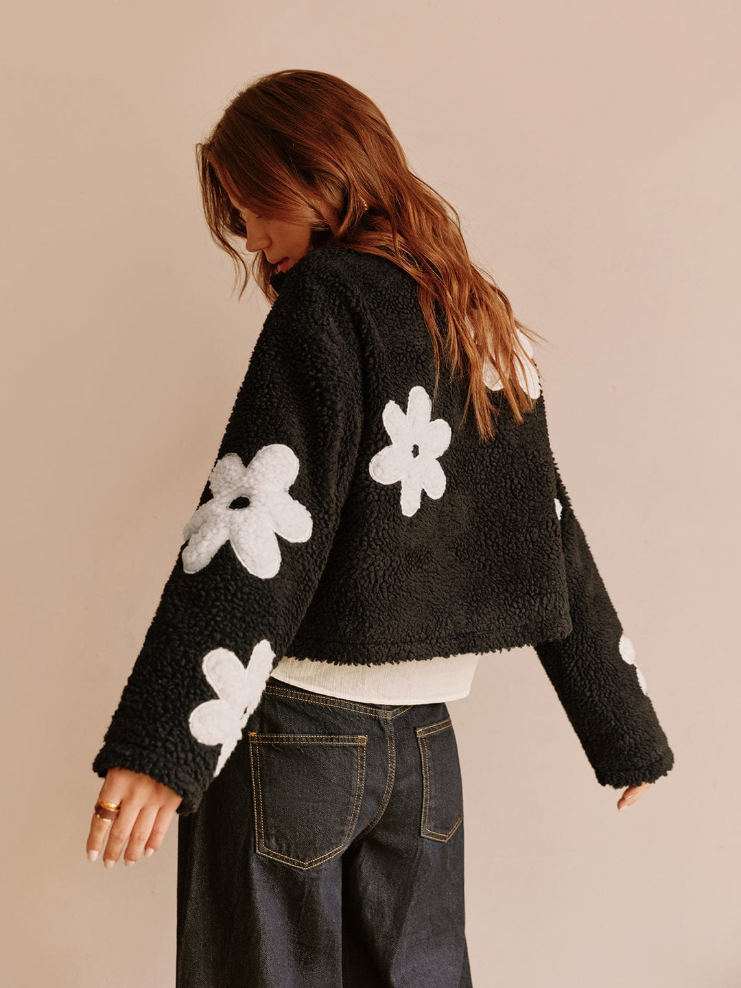 Vue arrière de cette veste courte en fausse fourrure noire ornée de fleurs blanches, parfaite pour un look moderne et décontracté. Idéale pour une tenue confortable et tendance, cette veste se marie parfaitement avec un pantalon taille haute.
