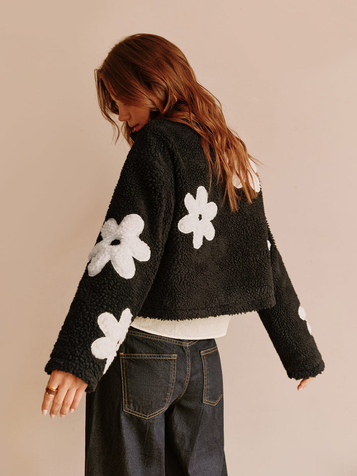 Vue arrière de cette veste courte en fausse fourrure noire ornée de fleurs blanches, parfaite pour un look moderne et décontracté. Idéale pour une tenue confortable et tendance, cette veste se marie parfaitement avec un pantalon taille haute.
