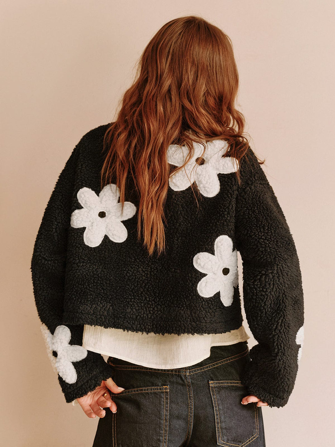 Vue arrière de cette veste courte en fausse fourrure noire avec des fleurs blanches. Son design moderne et confortable se marie parfaitement avec un pantalon taille haute, idéale pour un look tendance et décontracté.