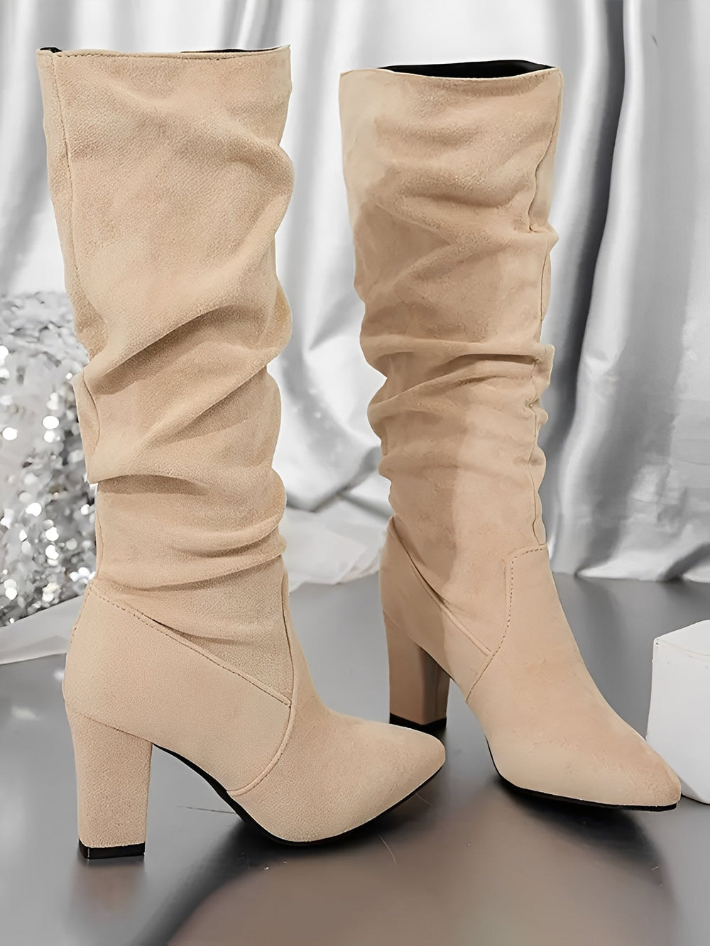 Paire de bottes hautes en daim beige à talons épais, avec une tige plissée et une silhouette élégante, présentées debout sur un sol brillant devant un fond satiné argenté.